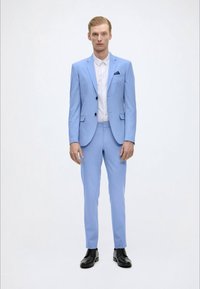Fato azul claro com um casaco de um botão, calças de corte slim, botões pretos e uma camisa branca. Sapatos pretos completam o look.