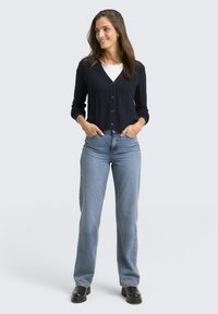 Marineblauer gerippter Cardigan mit fünf Knöpfen, kombiniert mit hellblauen Jeans im Straight-Leg-Schnitt und schwarzen klobigen Stiefeln, vor einem hellgrauen Hintergrund.