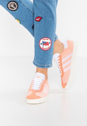 Piernas con vaqueros azules con parches y zapatillas Adidas en color durazno y blanco sobre un suelo blanco, con un pie ligeramente levantado.