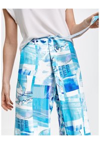 Kvinna iklädd byxor i blått och vitt med abstrakta mönster, knyter ett matchande tygbälte runt midjan, parat med en vit topp.