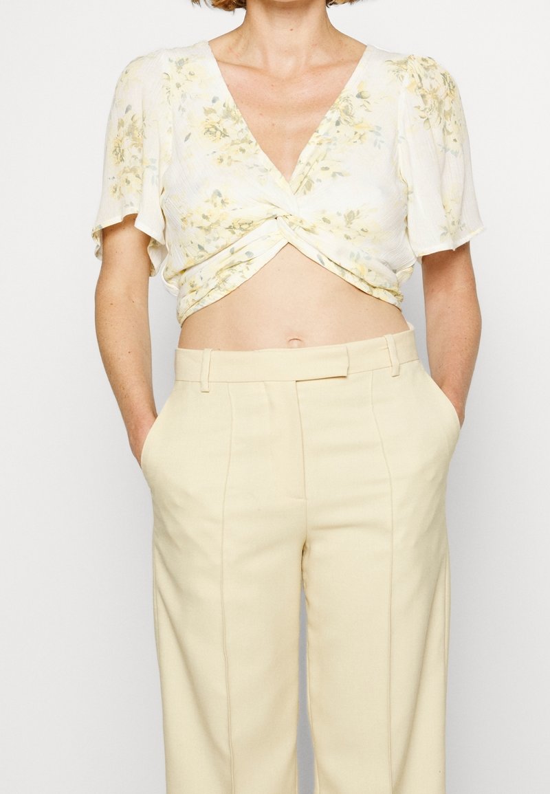 Weißes Blumen-Crop-Top mit V-Ausschnitt und verdrehtem Vorderteil, kombiniert mit hoch taillierten, hellbeigen Hose mit seitlichen Taschen und taillierter Passform.