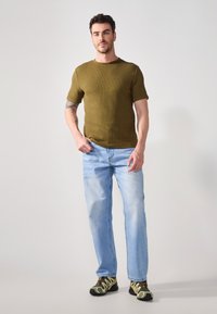 Cămașă scurtă cu mâneci scurte, de culoare verde măslin, blugi drepți de culoare albastru deschis și pantofi sport negri-verzi cu accente galbene.