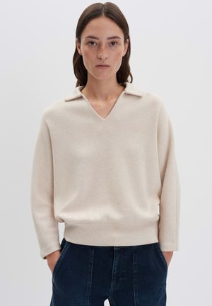 Vrouw met stijlbruin haar, gekleed in een crèmekleurige langemouwen trui met een kraag en donkerblauwe hoge taillebroek, handen in haar zakken.