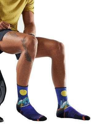 Persona con gambe tatuate che indossa calzini blu con un motivo paesaggistico, seduta con le ginocchia piegate contro uno sfondo bianco.