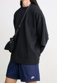 Person som bär en svart oversized sweatshirt, marinblå Nike-shorts och en liten svart crossbody-väska mot en helt vit bakgrund.