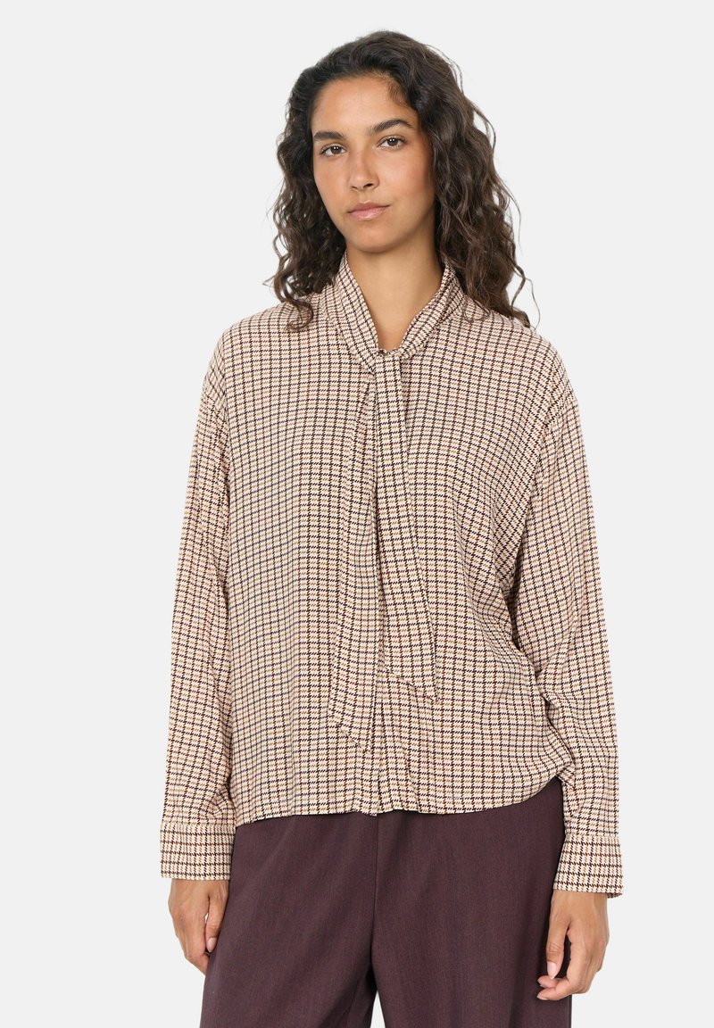 Femme aux cheveux bouclés et foncés portant un chemisier beige et marron à carreaux à manches longues et un pantalon marron foncé, debout devant un fond uni.