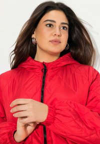 Red quilted jacket with a hood, featuring a front zipper, elastic cuffs, and smooth texture. Model holds hands near the zipper.