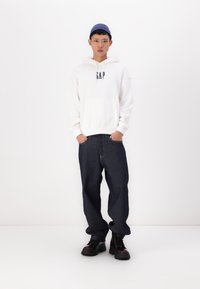 Sweat à capuche blanc avec le logo "GAP", jean bleu foncé et bottes noires. Le modèle porte un bonnet en tricot bleu. L'arrière-plan est blanc uni.