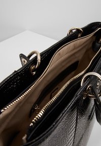 Sac à main en cuir verni noir avec une surface texturée, une quincaillerie dorée et un intérieur beige comprenant plusieurs poches et une fermeture éclair.