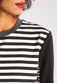Gestreifter Pullover in Schwarz und Weiß mit einem grauen Ausschnitt. Der Stoff ist weich und hat eine glatte Textur, mit horizontalen Streifen und einer kontrastierenden einfarbigen Fläche.