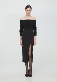 Top in maglia nera a spalle scoperte abbinato a una gonna nera con spacco alto. Il modello indossa collant trasparenti neri e décolleté nere decorate.