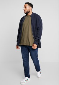 Marineblauwe geribde bomberjack over een olijfgroen t-shirt, gecombineerd met donkere spijkerbroek en witte sneakers met rode en groene strepen.