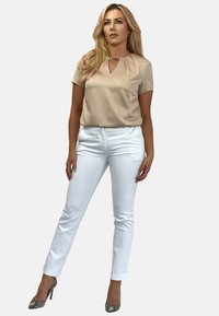 Gładka beżowa bluzka z detalem w postaci wycięcia, zestawiona z dopasowanymi białymi spodniami. Modelka nosi wzorzyste szpilki, prezentując nowoczesny design.