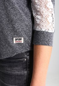Grauer Pullover mit Spitze an den Ärmeln, mit einem "Superdry Sportswear"-Label auf der Vorderseite. Kombiniert mit dunklen Jeans.