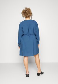 Vestido azul de mangas longas com um corte descontraído, cintura ajustada e detalhe de pregas nas costas. Pernas visíveis, combinado com mules de salto bloco pretos.