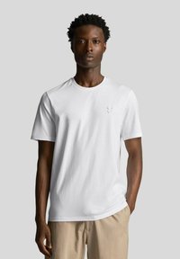 T-shirt bianca in cotone con colletto rotondo, maniche corte e un sottile dettaglio del logo ricamato sul lato sinistro del petto, abbinata a pantaloni beige.