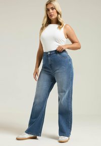 Vaaleansiniset leveät farkut, joissa on sivustudiot, korkea vyötärö ja etutaskut, yhdistettynä valkoiseen hihattomaan paitaan ja valkoisiin lenkkareihin.