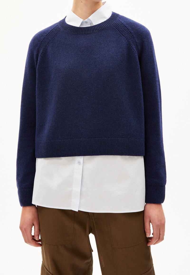 Marineblauwe cropped gebreide trui met geribbelde manchetten en halslijn, met een gladde textuur en raglanmouwen, gedragen over een wit overhemd.