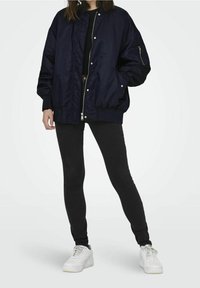 Námořnická bombera s předním zipem, elastickými manžetami a bočními kapsami, v kombinaci s černými legínami a bílými teniskami.