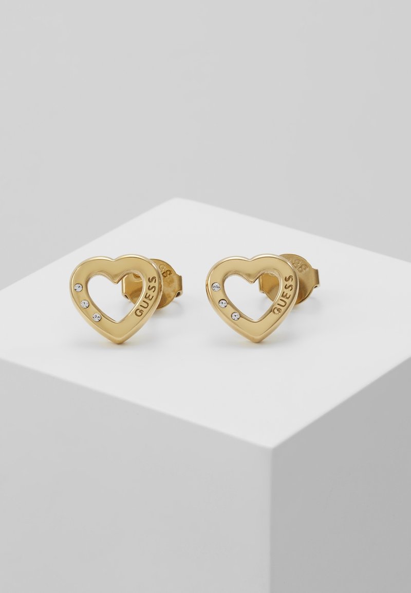 Boucles d’oreilles clous en forme de cœur en or avec trois petits cristaux et le mot "GUESS" gravé sur le côté, exposées sur une plateforme blanche.