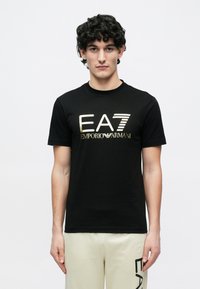 Fiatal férfi göndör hajjal, fekete EA7 Emporio Armani pólót és bézs nadrágot visel, melyen az EA7 logó látható a combnál, fehér háttér előtt áll.