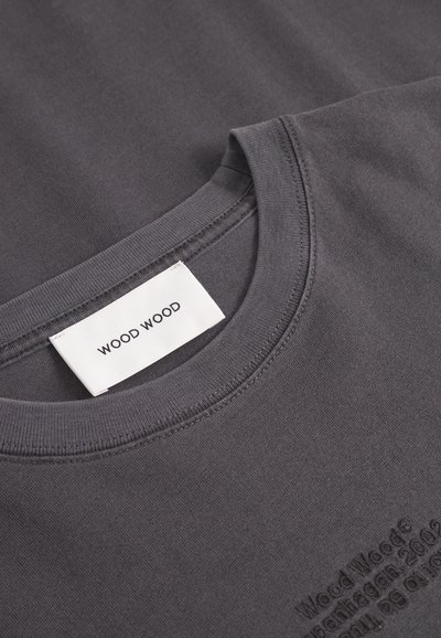 Prim-plan al unui sweatshirt negru închis cu guler rotund, având eticheta albă Wood Wood și un text subtil embossat pe țesătură.
