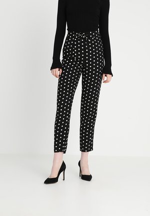 Femme portant un pantalon noir taille haute à pois blancs, un haut noir à manches longues et des chaussures noires à talons hauts, debout sur un fond uni.
