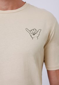 Beige cotton t-shirt featuring a black graphic of a hand making the "shaka" sign on the left chest area, with a classic round neck.