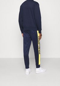 Marineblauer Trainingsanzug mit gelbem Seitenstreifen und "POLO SPORT"-Logo. Weicher Stoff, lässige Passform und passgenaue marineblaue Jogginghosen mit Bündchen. Weiße Sneakers.