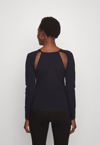 Svart topp med långa ärmar som har utskärningsdetaljer på baksidan och en kroppsnära passform. Tillverkad av mjukt tyg med en slät yta.