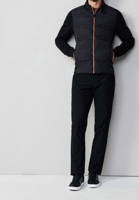 Homme portant un gilet noir zippé avec une fermeture éclair orange, une chemise à manches longues noire, un pantalon noir et des baskets noires à semelles blanches, debout contre un mur.