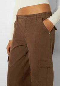 Bruine cargobroek met knoopsluiting, zijzakken en een elastische tailleband. De stof heeft een gladde textuur met verstevigde naden.