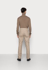 Maglione a collo alto marrone abbinato a pantaloni beige, caratterizzato da una vestibilità rilassata e tasche posteriori, indossato con scarpe nere.