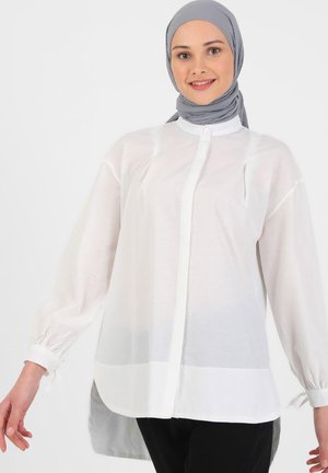 White shirt with long sleeves, soft fabric, button-up front, and two chest pockets. Elastic cuffs and a longer back hem.