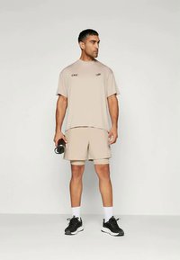 Μπεζ αθλητικό μπλουζάκι με λογότυπο και ταιριαστά σορτς. Το μοντέλο κρατάει ένα μπουκάλι νερού. Τα παπούτσια είναι μαύρα με λευκές λεπτομέρειες.