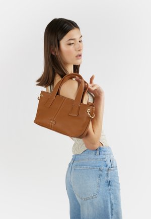 Donna con top senza maniche e jeans che tiene una borsa in pelle marrone sulla spalla su sfondo bianco.