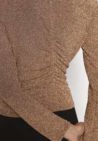 Top a maniche lunghe in tessuto dorato lucido con una finitura texture e brillante. Presenta dettagli arricciati lungo i lati e maniche aderenti.