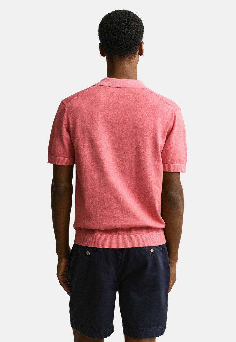 Homme debout, dos à la caméra, portant un t-shirt rose en maille à manches courtes et un short bleu marine foncé, sur un fond clair uni.