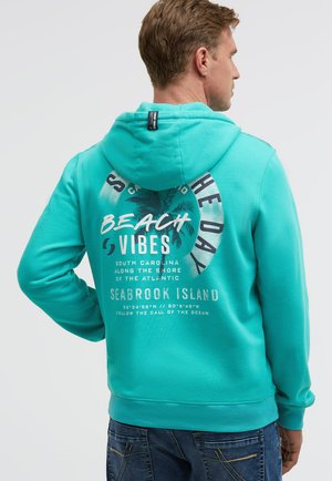 Man wearing turquoise hoodie with palm tree and text "Beach Vibes, Seabrook Island, South Carolina" standing against light background.