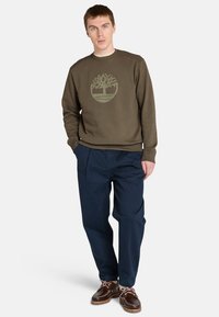 Sudadera verde oliva con un logo de árbol circular, combinada con pantalones sueltos de ajuste azul marino y zapatos marrones con cordones blancos.