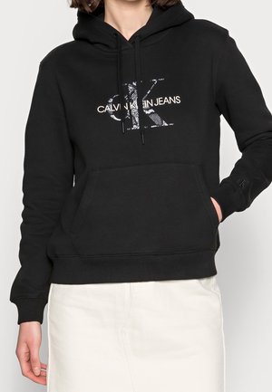 Kvinna i en svart huvtröja med "Calvin Klein Jeans"-logotyp, stående med en hand i fickan, tillsammans med en ljusfärgad kjol.