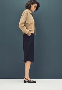 Chaqueta de ante beige con un diseño corto y un dobladillo acanalado, combinada con una falda negra hasta la rodilla y tacones estilo slingback negros.