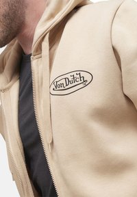 Sweatshirt zippé à capuche beige avec un logo noir "Von Dutch" sur la poitrine. Matériau doux avec une texture lisse et cordons de serrage intégrés.