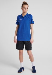Blå piké med kort ärm, med vit logotyp och chevrondetaljer. Kombinerad med svarta shorts och blå sneakers.