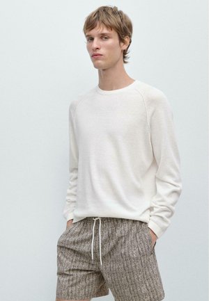 Młody mężczyzna z krótkimi, potarganymi włosami, ubrany w białą koszulę z długim rękawem i wzorzyste szorty ze ściągaczem, ręce w kieszeniach, na jednolitym tle.