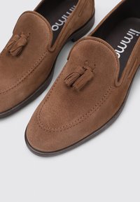Mocasines de ante marrón con suela plana, que presentan un acento de borlas y detalles de costura en una superficie suave. Logo embossado visible en el interior.