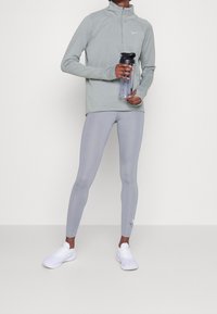 Lichtgrijs sporttop met lange mouwen en een halve rits, gecombineerd met lichtgrijze leggings en witte sneakers. Waterfles in hand.