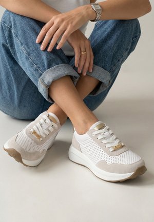 Osoba siedząca po turecku, ubrana w niebieskie podwinięte dżinsy oraz białe trampki z beżowymi akcentami i złotymi detalami.