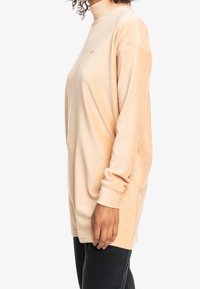 Sudadera de gran tamaño en color durazno, tejido suave, cuello alto, mangas largas, aberturas en los laterales, detalle sutil del logo cerca del pecho, corte relajado.