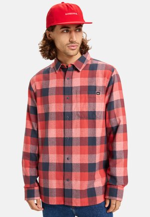 Rød og marineblå rutet flannel skjorte med lange ærmer, knapfront og en brystlomme. Båret med en ensfarvet rød kasket.